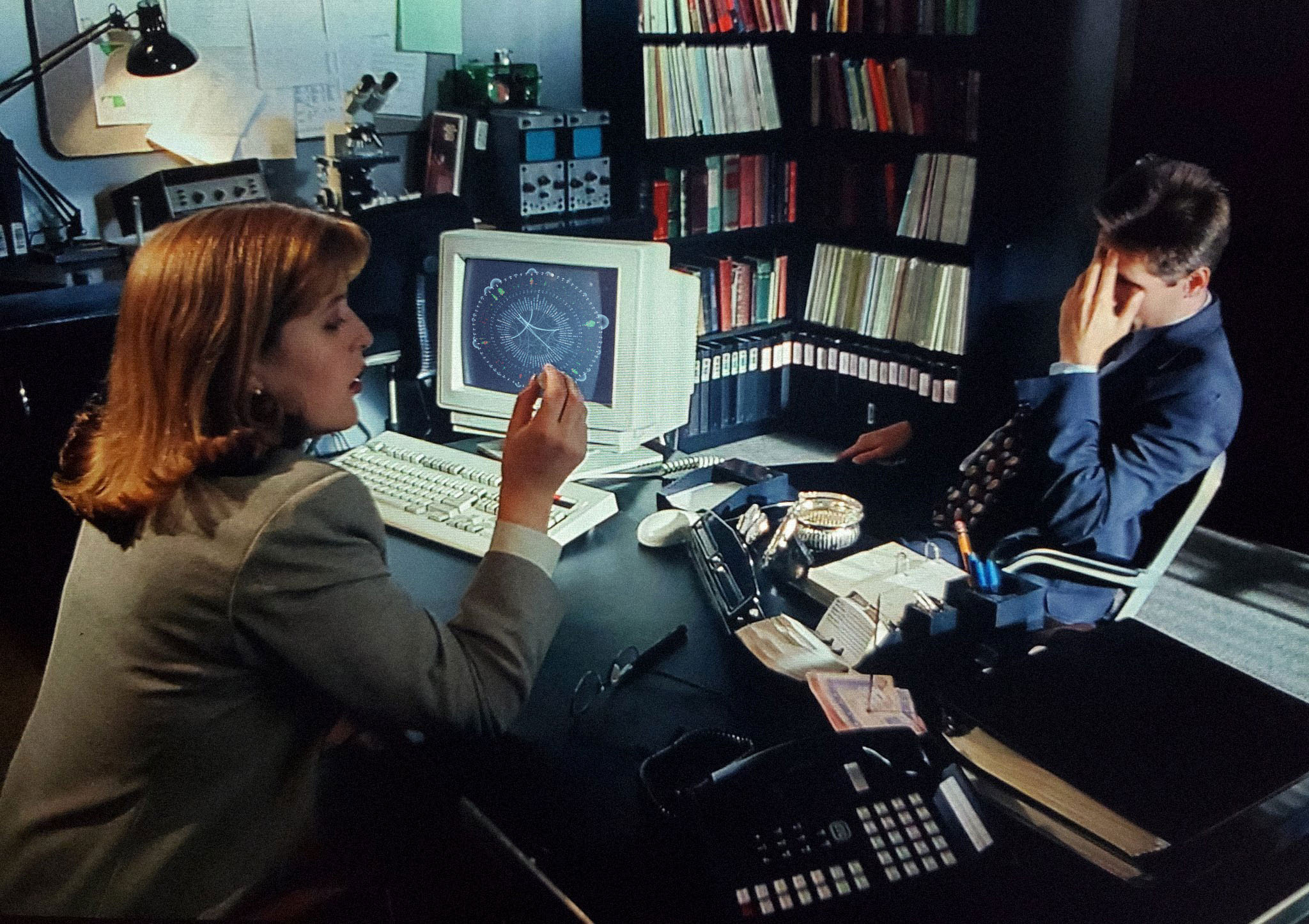 FBI agents Fox Mulder and Dana Scully are sitting in an office, with the data visualization 'Every Writer of Chris Carter's TV Shows' open on the monitor screen on the desk